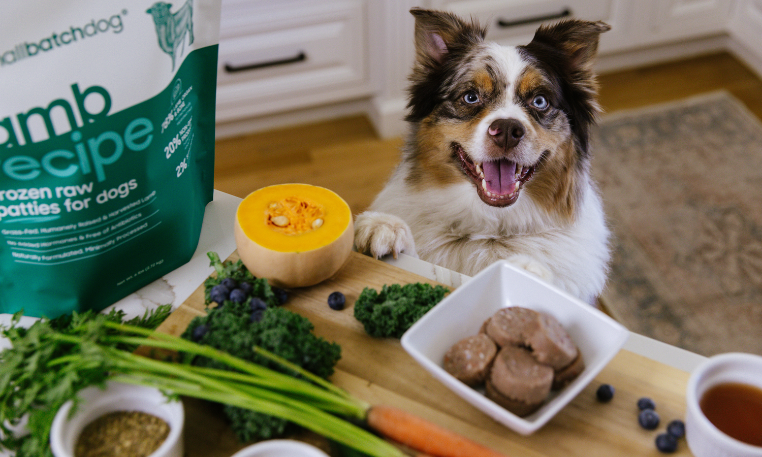 Dog with paws on counter featuring various vegetables and frozen raw patties.