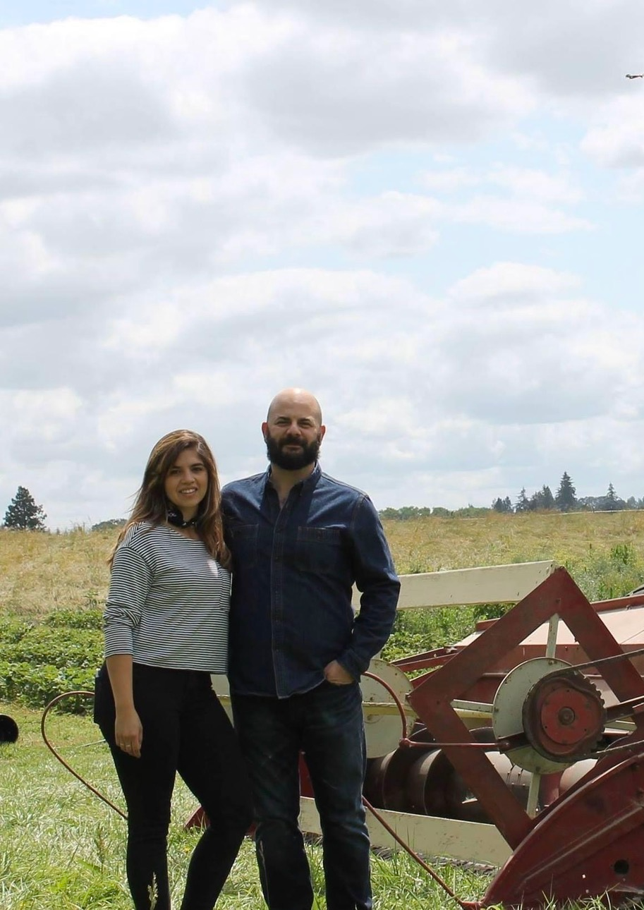 A man and a woman stand outdoors in a field next to a large vintage red combine harvester under a cloudy sky.