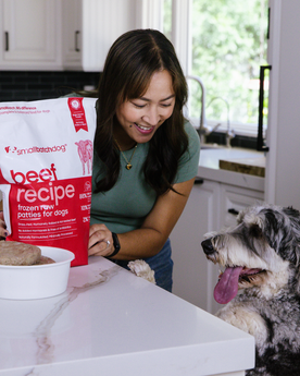 Woman looking at dog with paws on counter while holding a bag of frozen patties. 