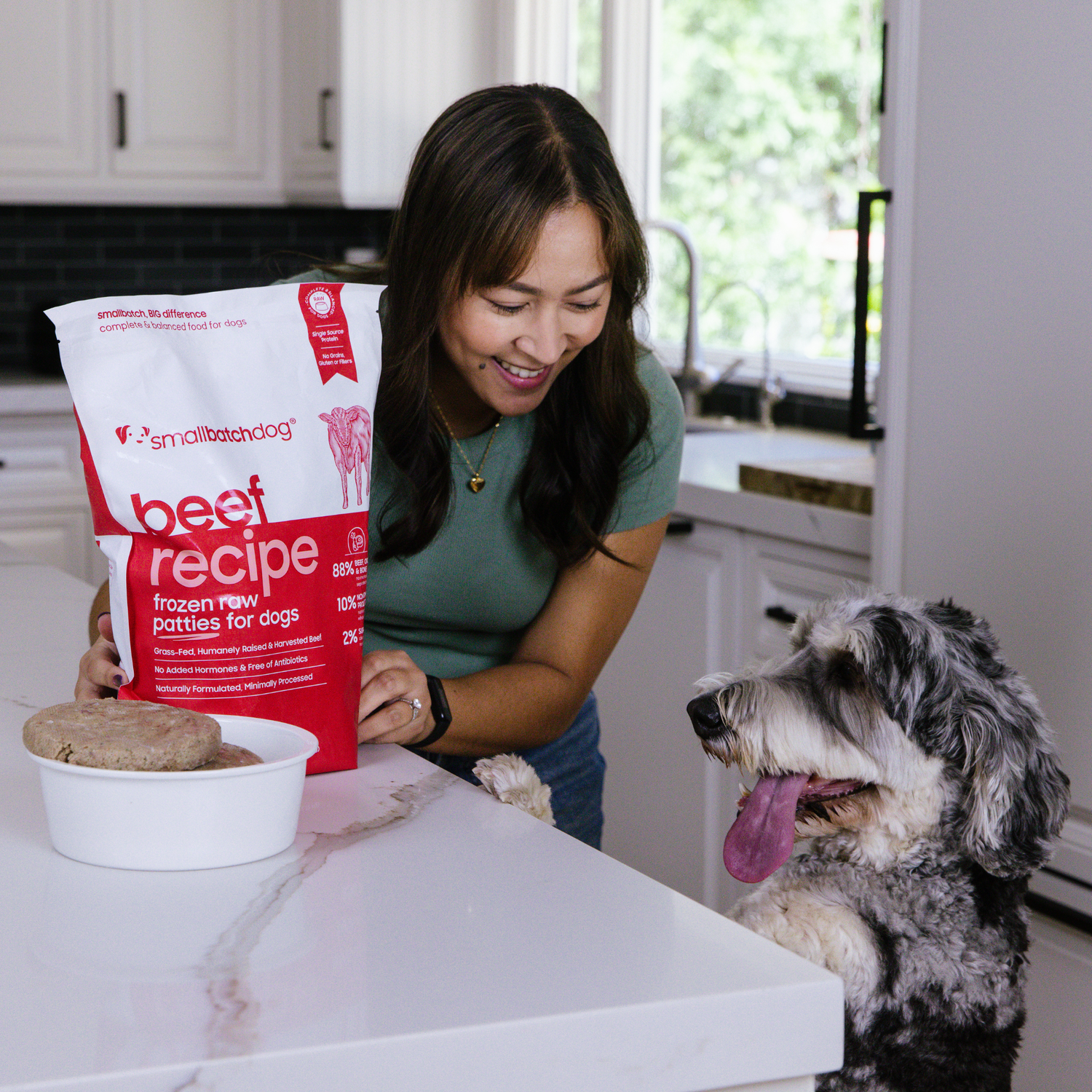 Woman holding a bag of smallbatch beef frozen raw dog food with a dog in a kitchen.